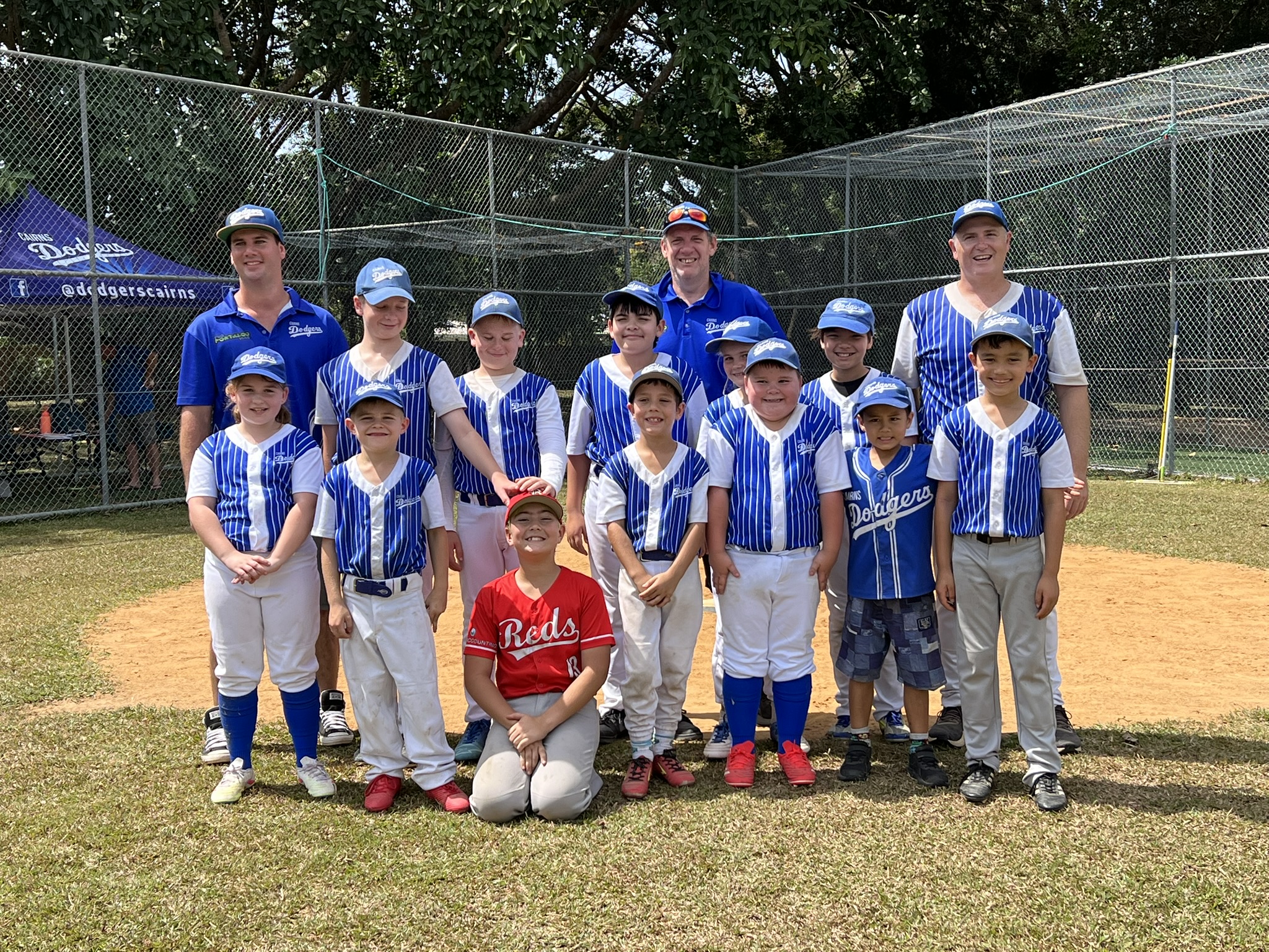 Group of kids and coaches in blue and white baseball uniforms posing for a team photo on a sunny field behind a chain-link fence.