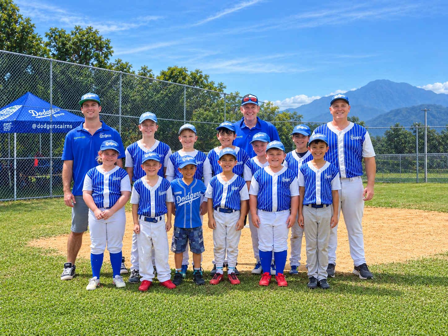 Youth baseball team in blue and white uniforms posing with coaches on a sunny field, mountains in the background.