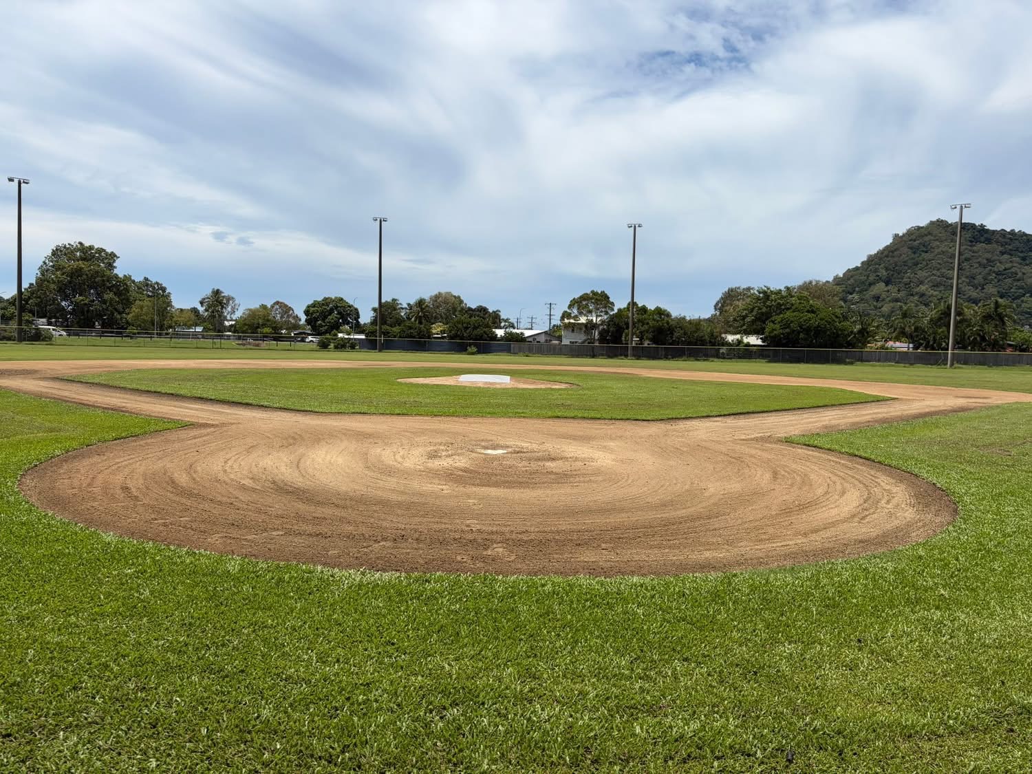 A baseball field with a dirt infield, green grass, and a pitched pitcher's mound under a blue sky.