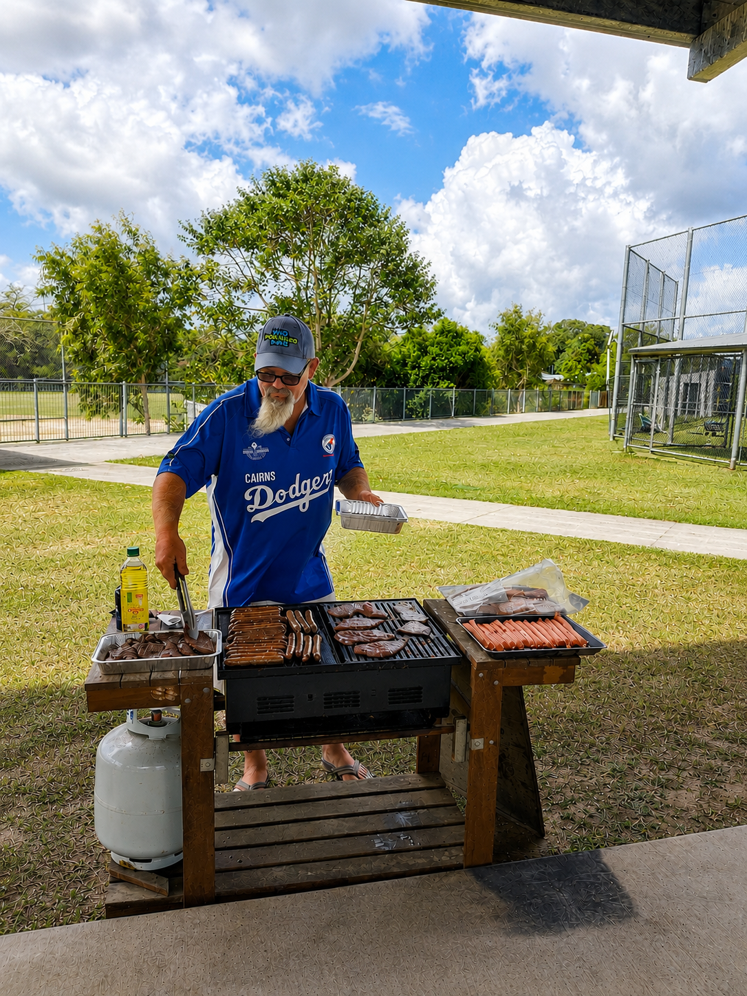 Man in a blue Cairns Dodgers shirt grills sausages on an outdoor barbecue, with a propane tank and trays of meat nearby in a park setting.