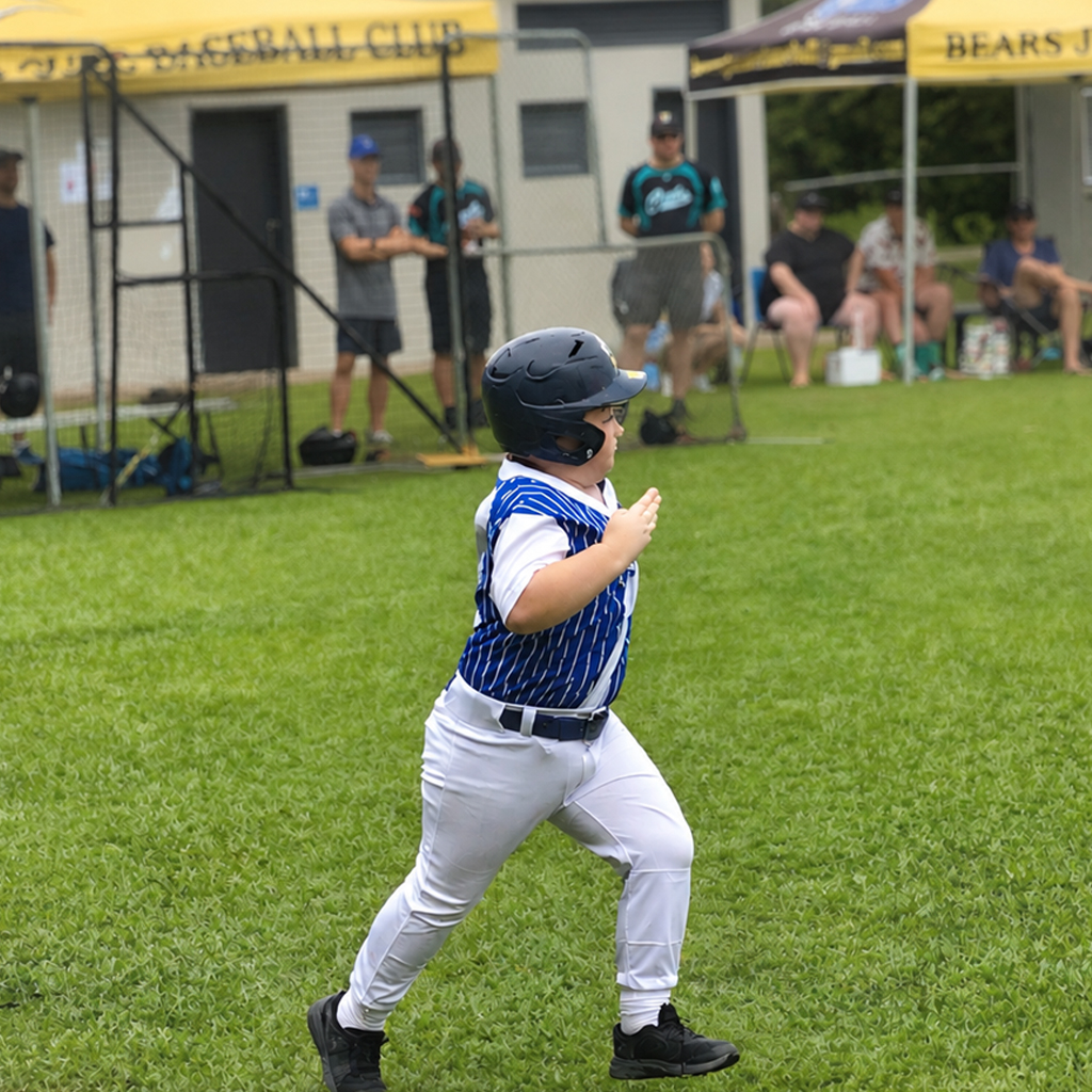 Young baseball player in a blue striped jersey runs on the grass field, wearing a helmet, with spectators under tents in the background.