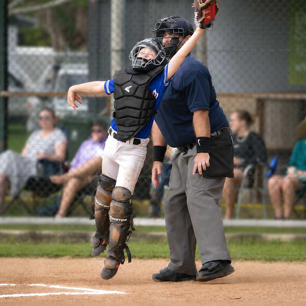 Catcher in blue, chest protector and helmet leaps at home plate while an umpire stands nearby and spectators watch behind a chain-link fence.