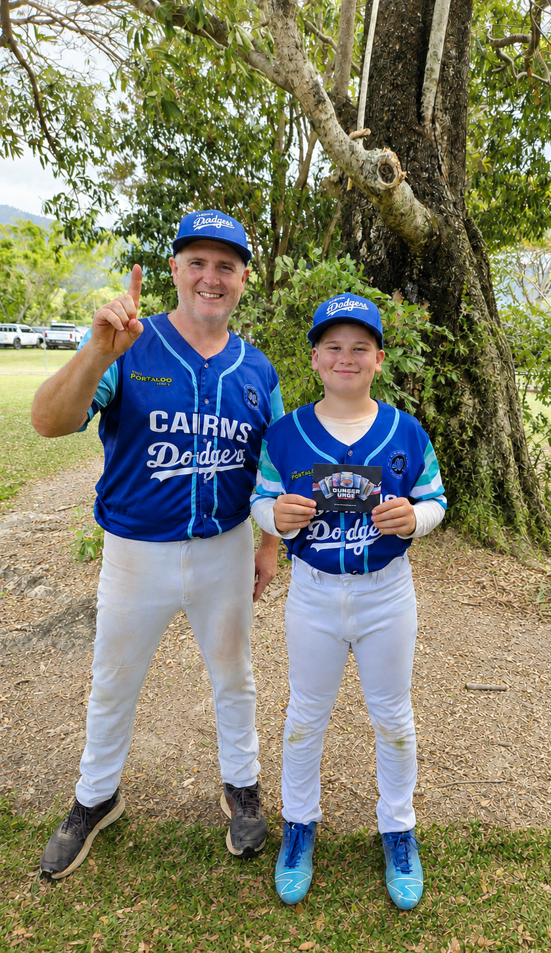 Two baseball players in blue Dodgers/Cairns uniforms pose outdoors under a tree; one man lifts a finger in greeting and the other girl holds a card.