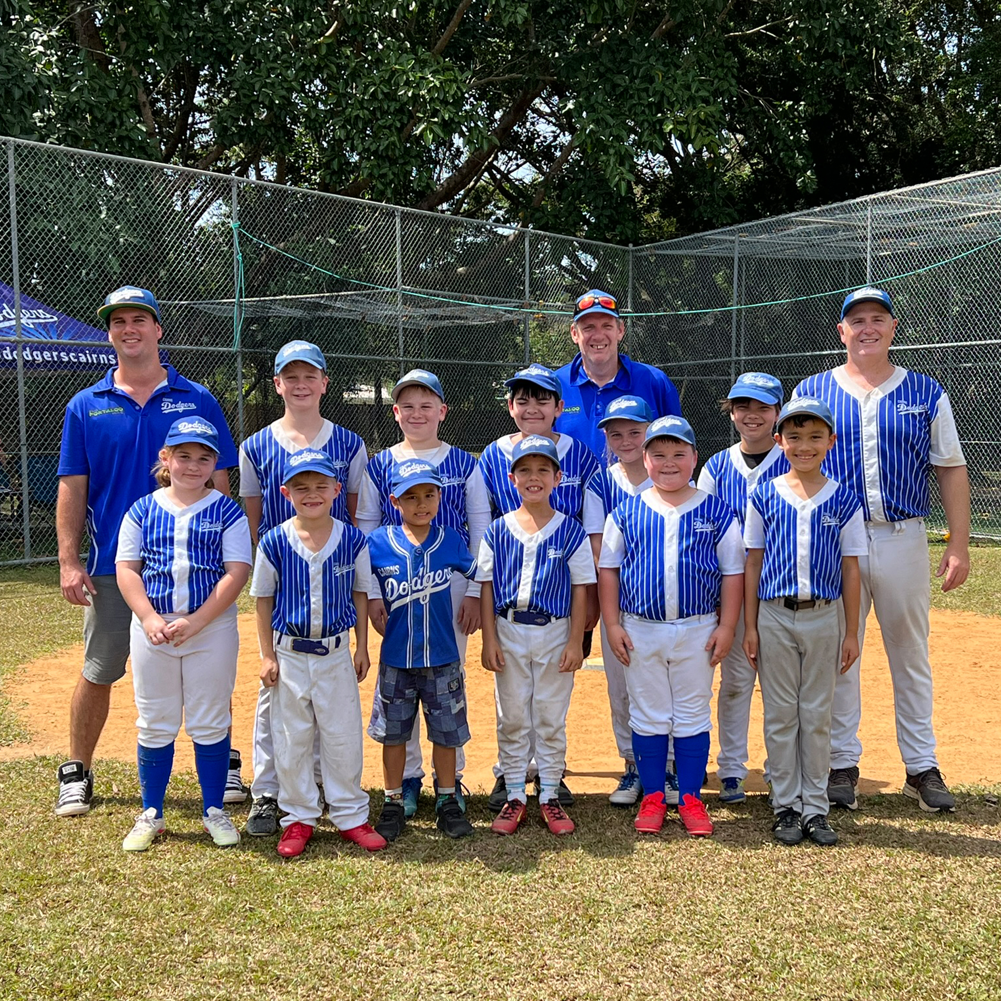 Group of children in blue and white striped uniforms posing for a team photo on a dirt field with a chain‑link fence in the background.